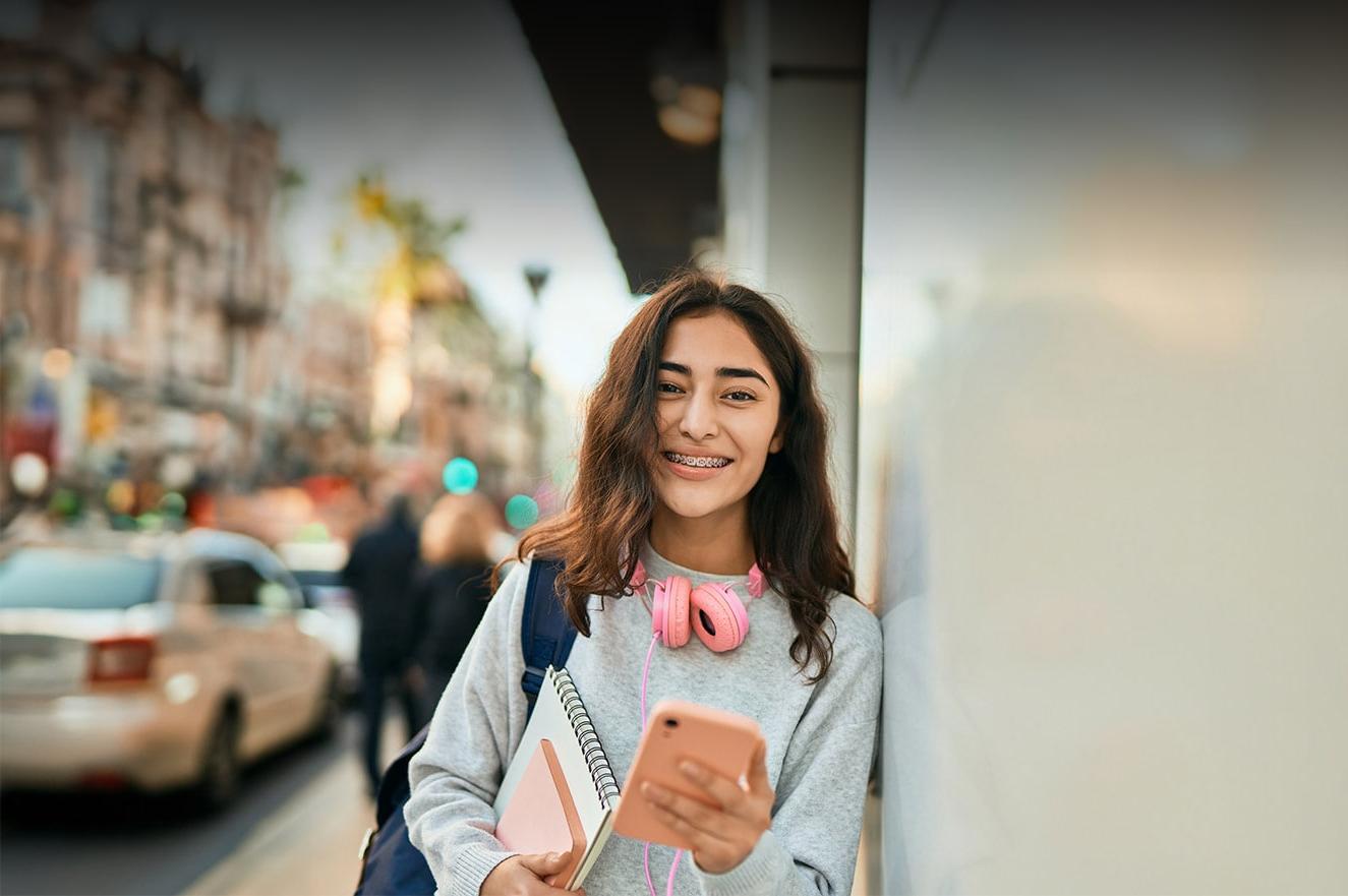 An online school student leaning against a wall and holding a pink phone and a notepad